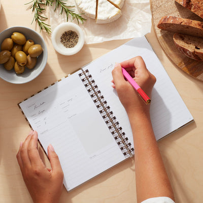 A person writing in a lemon recipe journal, surrounded by olives, bread, and cheese, showcasing culinary creativity.