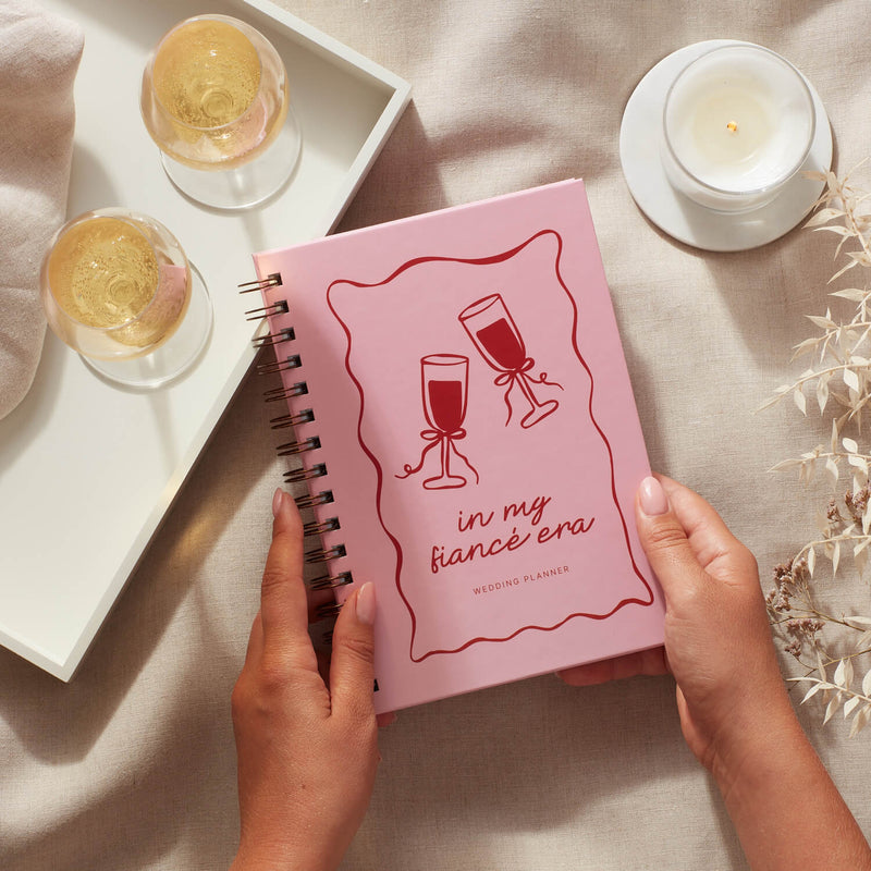 Hands holding a pink wedding planner titled 'in my fiancé era' with wine glasses illustration and graceful typography.