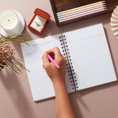 Person writing in a wedding planner book, focusing on a wedding guest list with soft pastels and modern design.