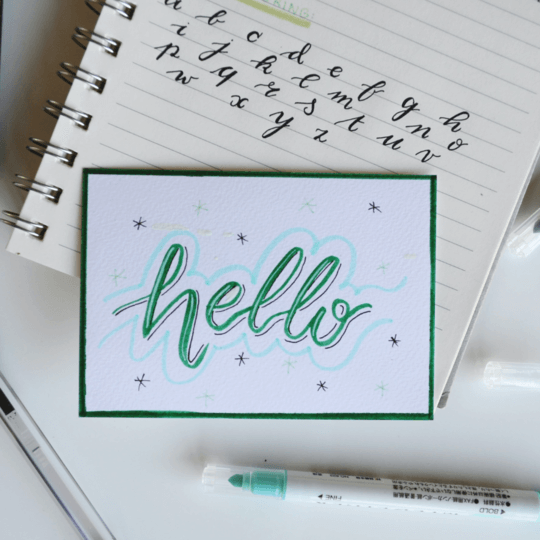 Image of word hello written out on card with notebook behind with alphabet