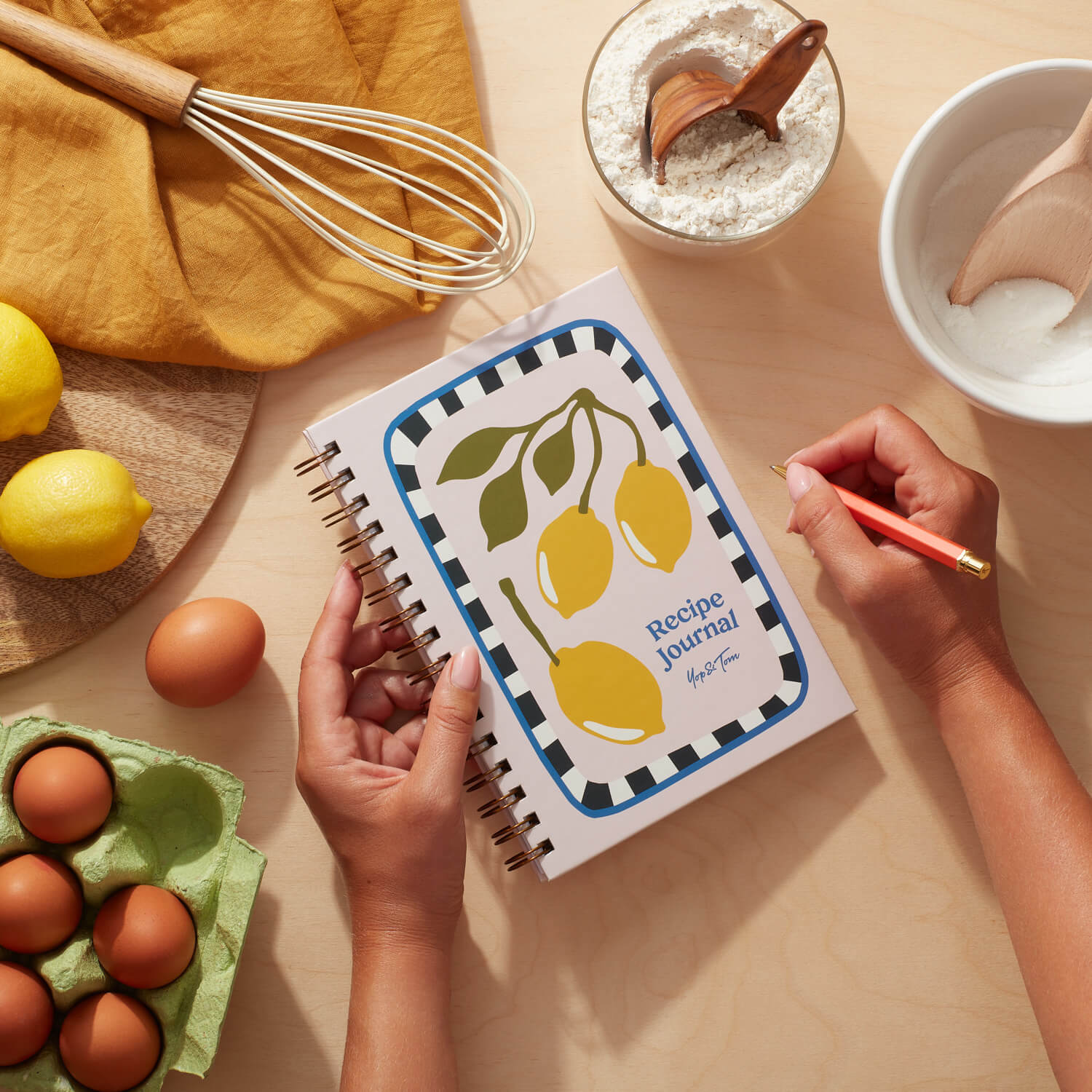 Lemon recipe journal with vibrant lemon design on a kitchen table next to ingredients and a hand writing.