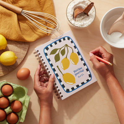 Lemon recipe journal with vibrant lemon design on a kitchen table next to ingredients and a hand writing.