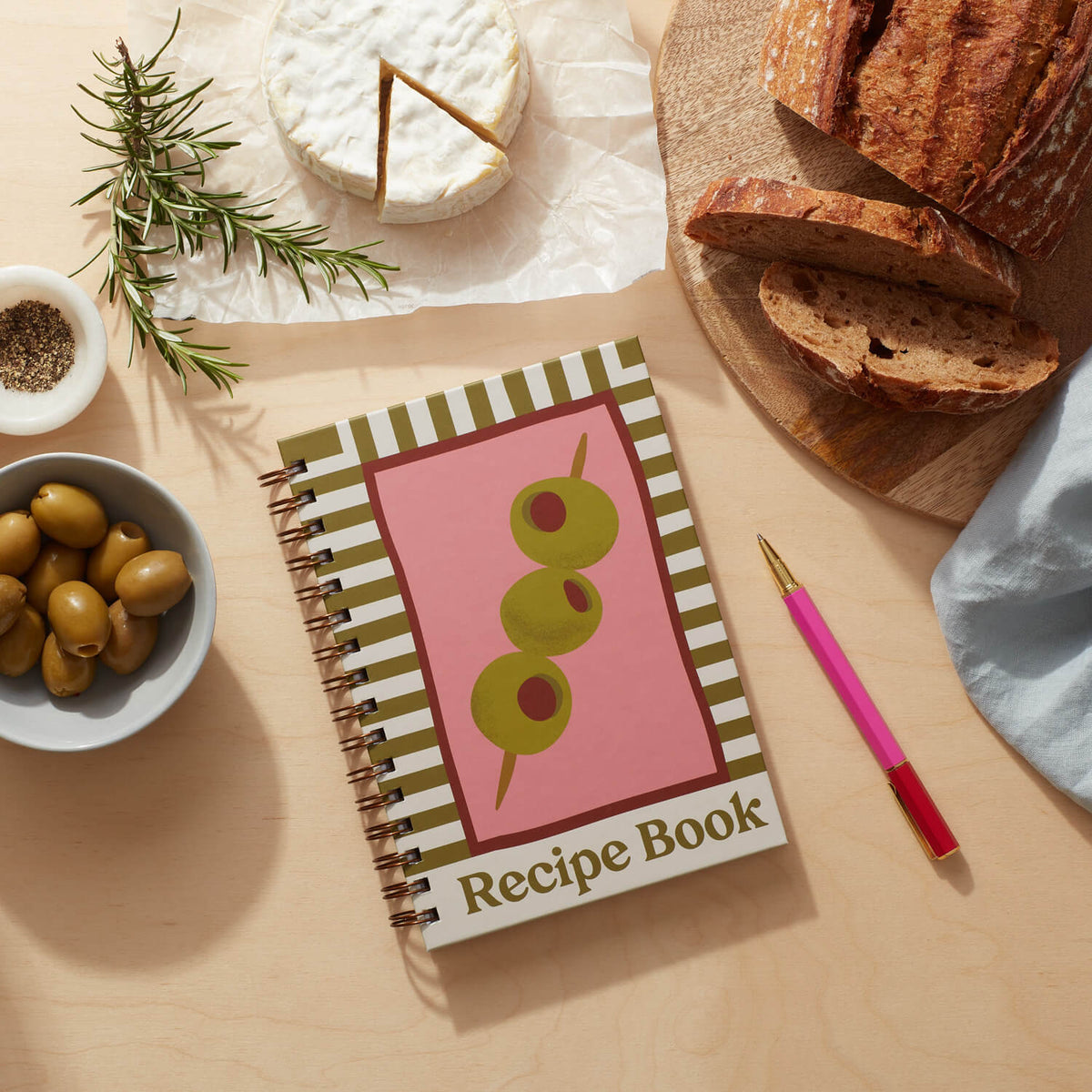 Recipe journal featuring an olive green cover with playful typography and culinary motifs, surrounded by bread and olives.