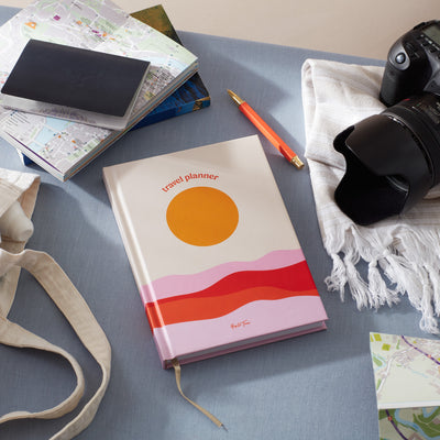 Travel planner with sunset design on a desk surrounded by maps and a camera.