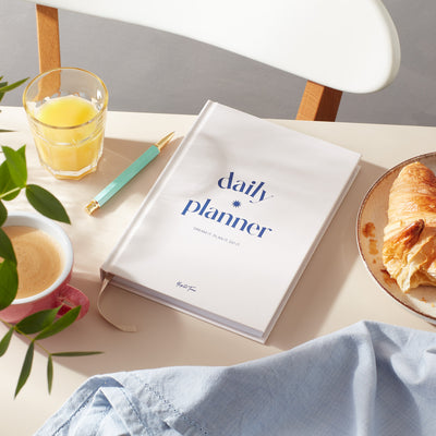 Daily planner on a desk with coffee, pastry, and juice, featuring a soft cover design for daily scheduling.